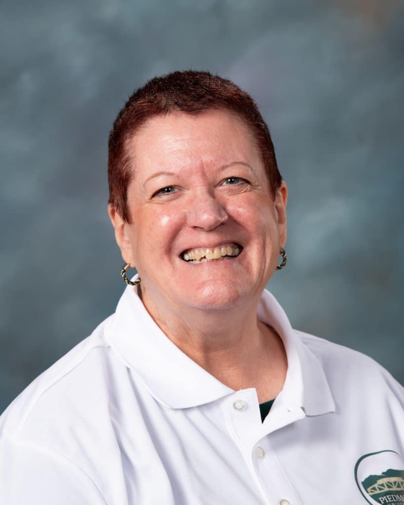 A smiling person with short brown hair, wearing a white collared shirt with a logo and gold earrings, poses in front of a blurred blue and gray background.