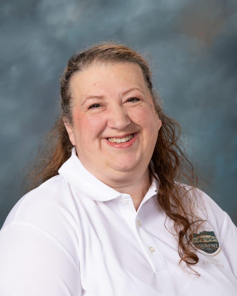 A woman with long, wavy brown hair smiles at the camera. She is wearing a white collared shirt with a green and gold embroidered logo on the chest, set against a soft blue-gray background.