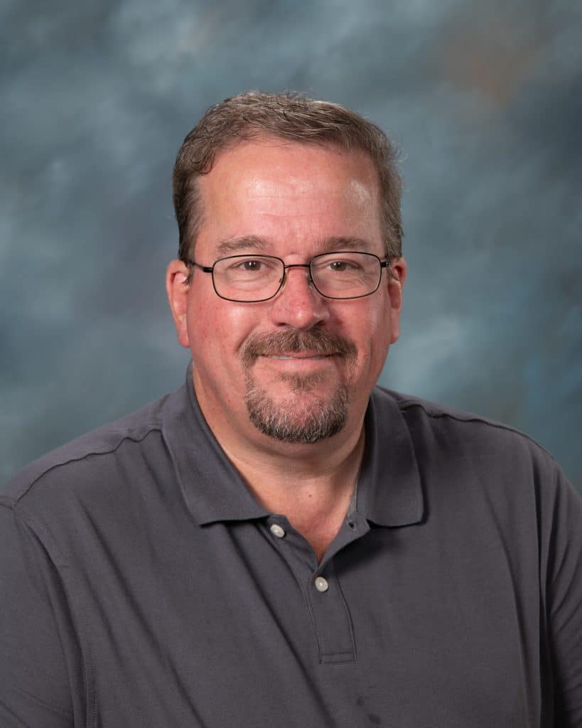 A man with short brown hair, glasses, and a goatee is smiling at the camera. He is wearing a dark gray polo shirt and is posed in front of a blue and gray mottled background.