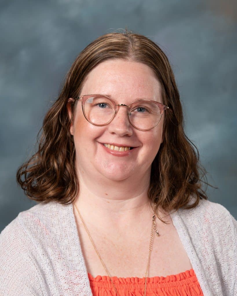 A woman with shoulder-length brown hair wearing glasses, a light gray cardigan, and an orange top smiles at the camera against a mottled blue-gray background.