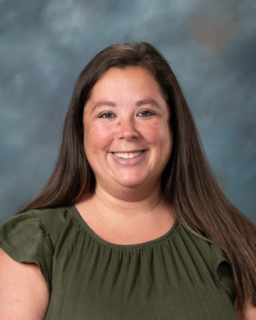 A woman with long brown hair wearing a short-sleeved olive green top smiles in front of a gray-blue mottled background.