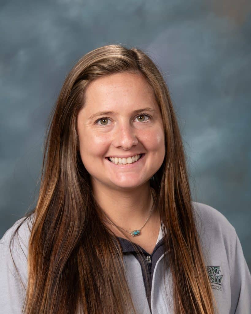 A woman with long brown hair smiling, wearing a light gray zip-up jacket and a turquoise necklace, posing in front of a blue-gray studio background.