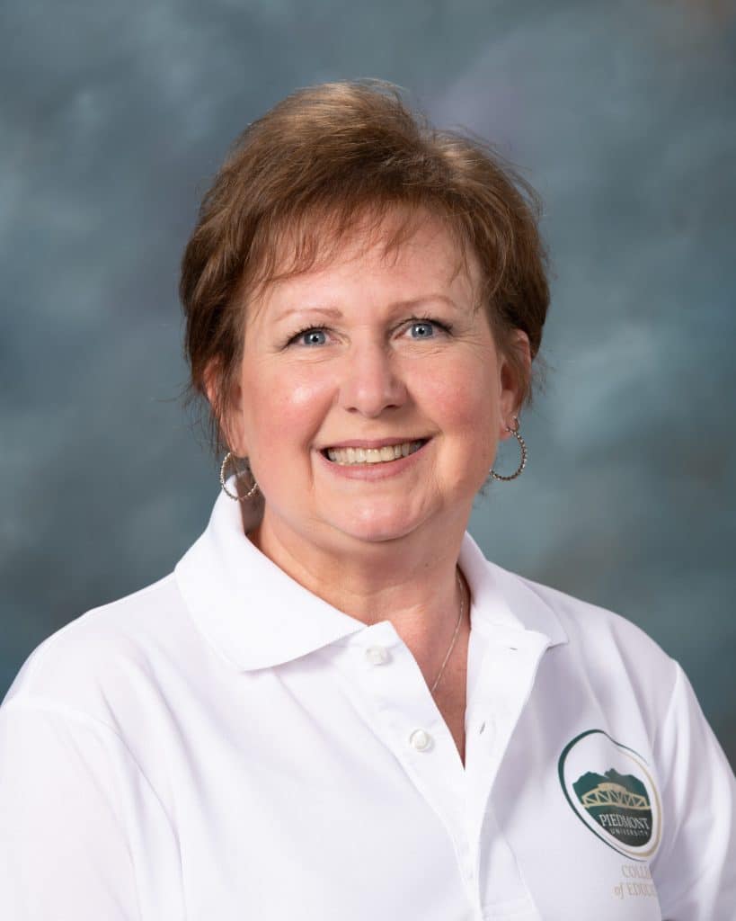 A woman with short light brown hair smiles at the camera. She is wearing hoop earrings and a white collared shirt with a Piedmont University College of Education logo. The background is a blue-gray gradient.