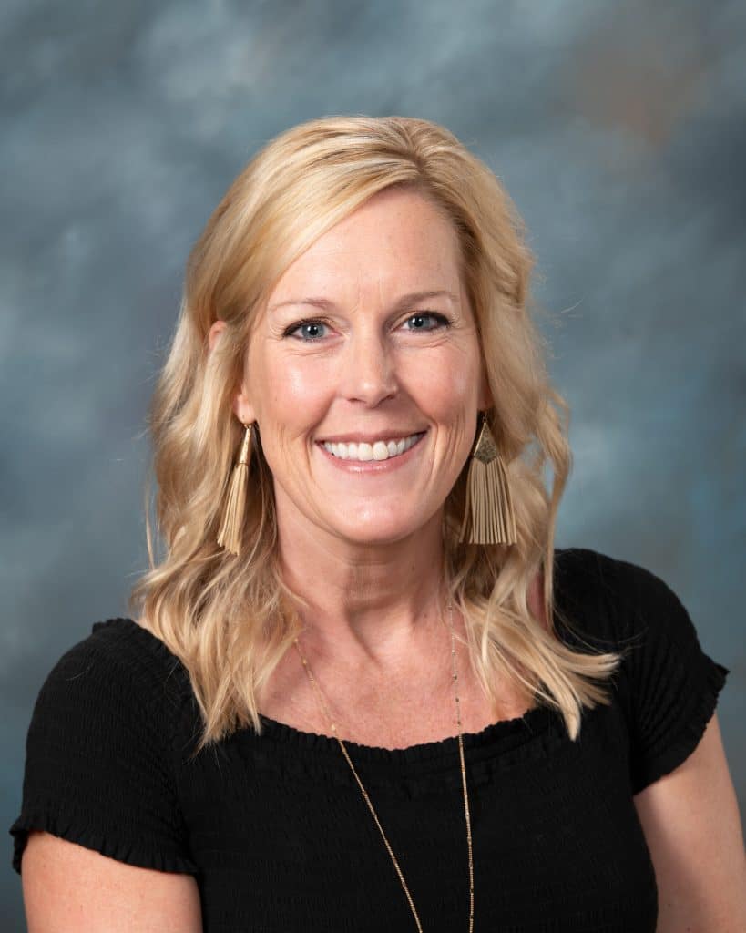 A woman with shoulder-length blonde hair, wearing a black top, long gold earrings, and a necklace, smiles at the camera in front of a blue and gray studio background.