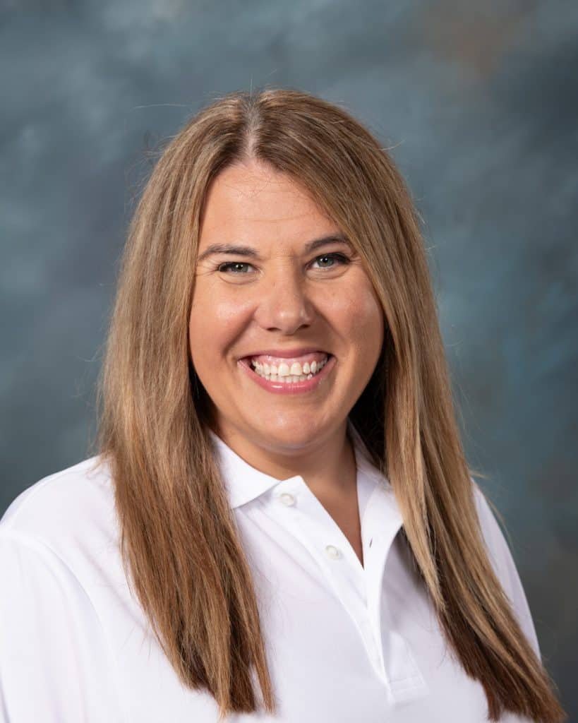 A woman with long straight light brown hair wearing a white collared shirt smiles at the camera. The background is a mottled blue and gray.