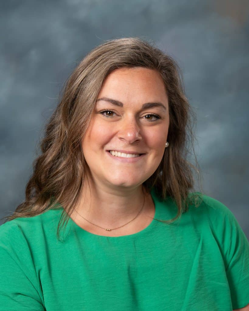 A woman with wavy brown hair wearing a green top and a subtle necklace, smiling at the camera against a gray, cloudy studio background.