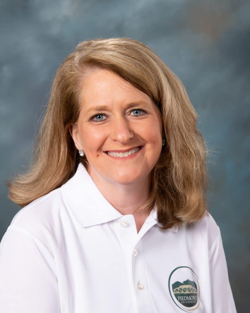 A woman with shoulder-length blonde hair and blue eyes smiles at the camera, wearing a white collared shirt with the Piedmont logo on it, set against a blue and gray studio background.