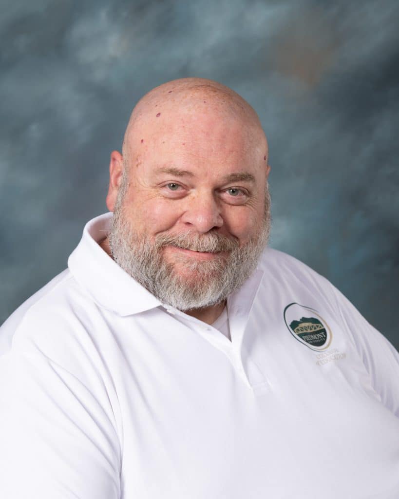 A man with a beard and bald head, wearing a white polo shirt with a green and gray logo, smiles at the camera against a blurred blue-gray background.
