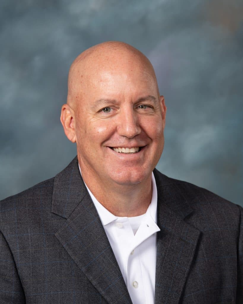 A smiling bald man in a dark checkered suit jacket and white collared shirt poses in front of a blurred gray-blue background.