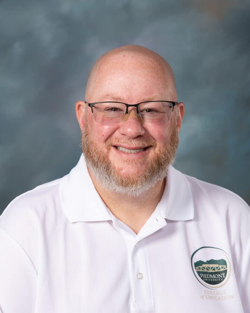 A smiling man with glasses, a short beard, and a bald head wears a white polo shirt with a Piedmont University College of Education logo, posing in front of a gray-blue background.