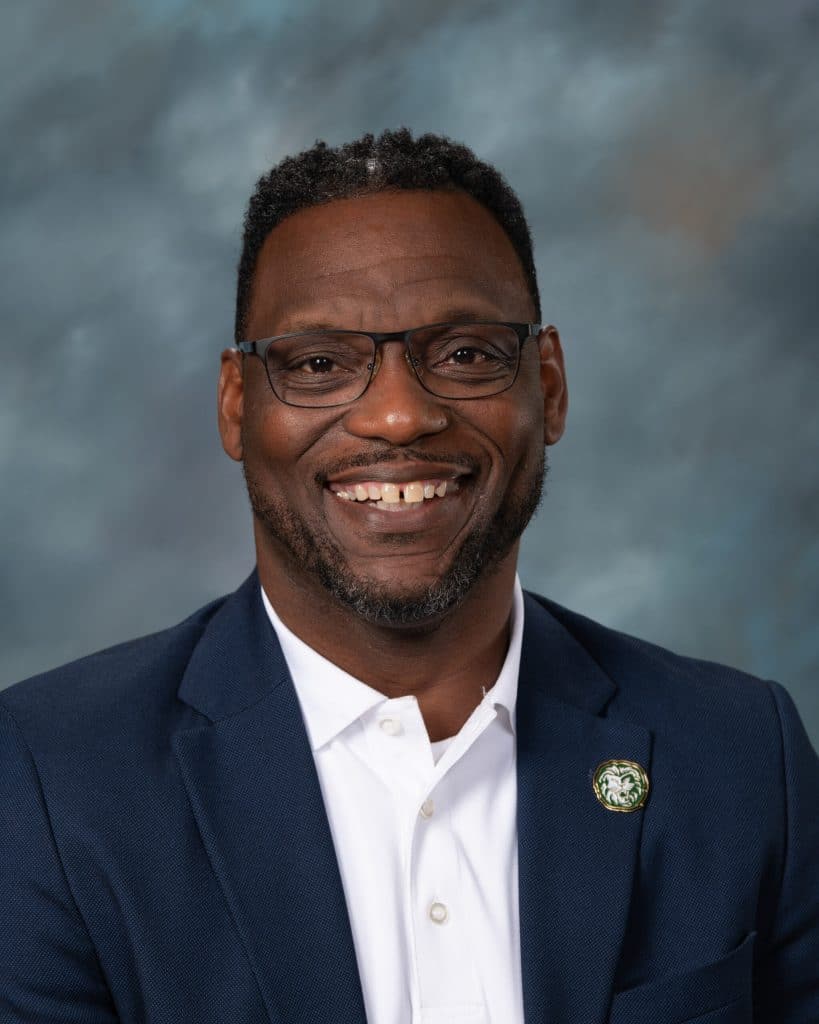 A smiling man with glasses and a short beard wears a navy blazer, white shirt, and a round lapel pin, posing in front of a blue-gray textured background.