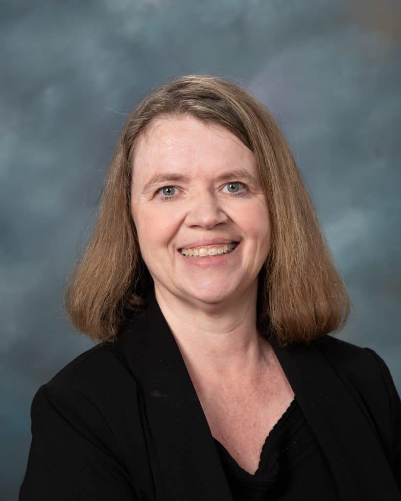 A woman with shoulder-length light brown hair smiles at the camera. She is wearing a black top and blazer, and is posed against a cloudy gray backdrop.