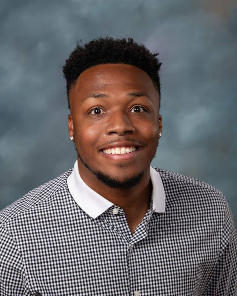 A smiling man with short curly hair and stud earrings, wearing a black and white checkered shirt with a white collar, posing in front of a blue and gray mottled background.