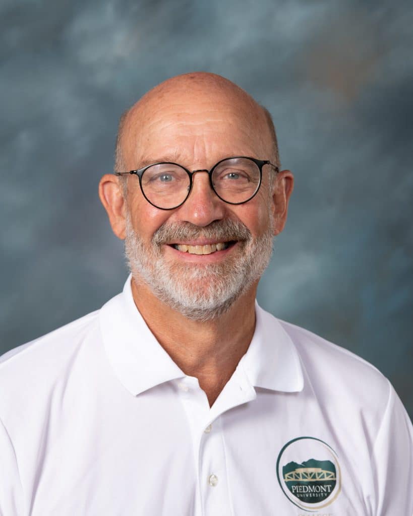 A smiling older man with glasses, a bald head, and a white beard wears a white polo shirt with a Piedmont logo, posing in front of a cloudy blue-gray background.
