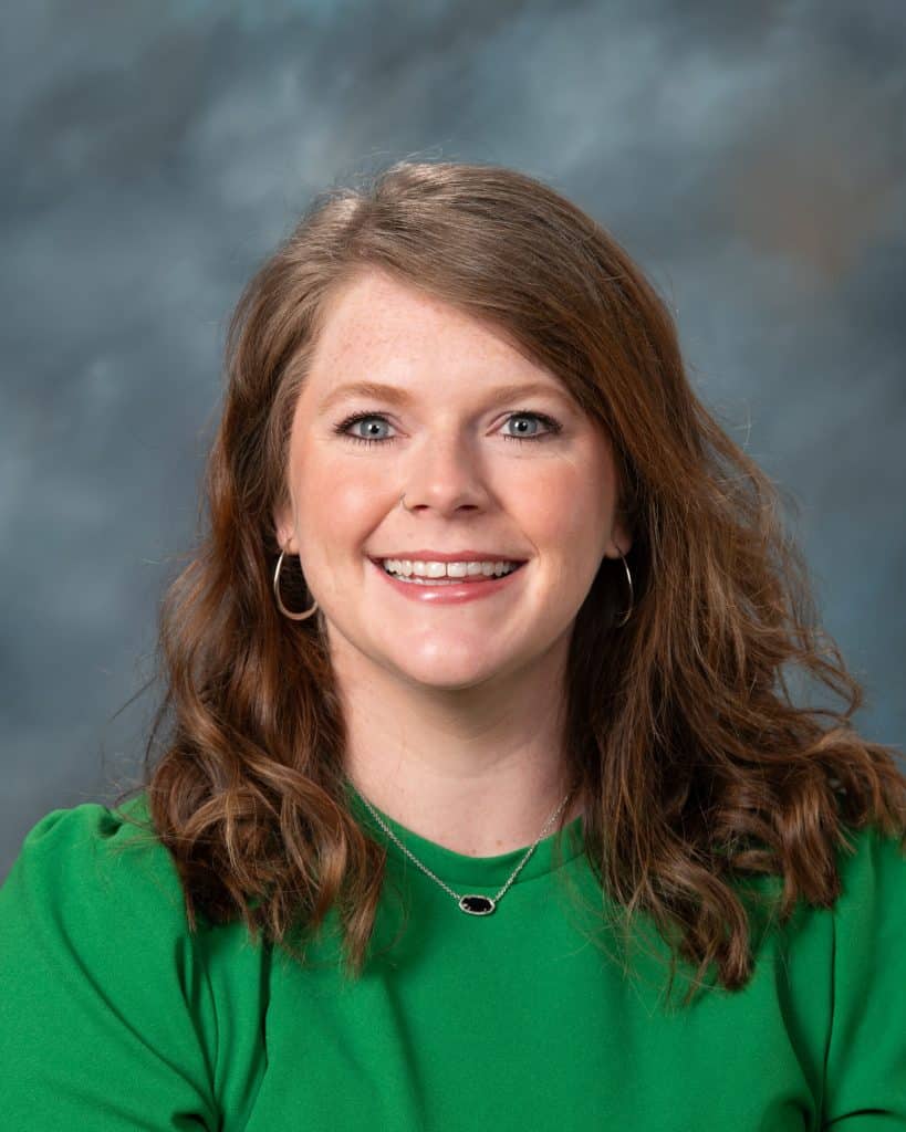 A woman with wavy brown hair, wearing a green top, hoop earrings, and a necklace, smiles at the camera in front of a gray-blue background.