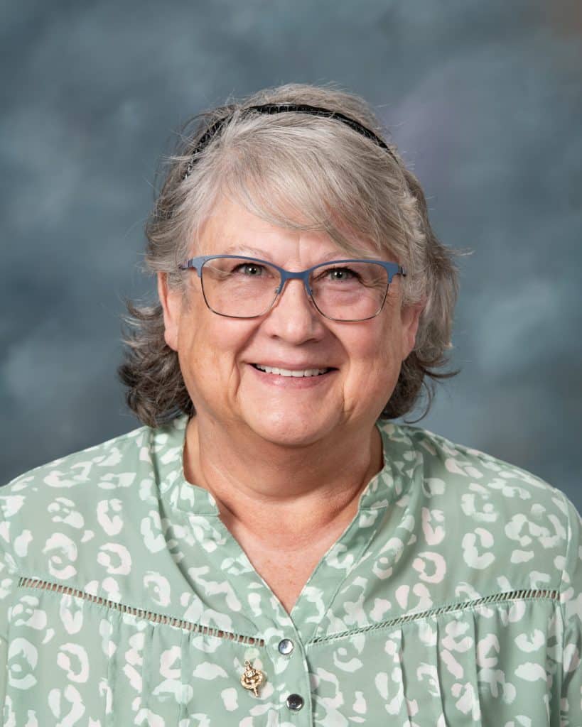 Smiling older woman with gray hair, wearing blue glasses and a mint green patterned blouse, poses in front of a soft, blue-gray background.