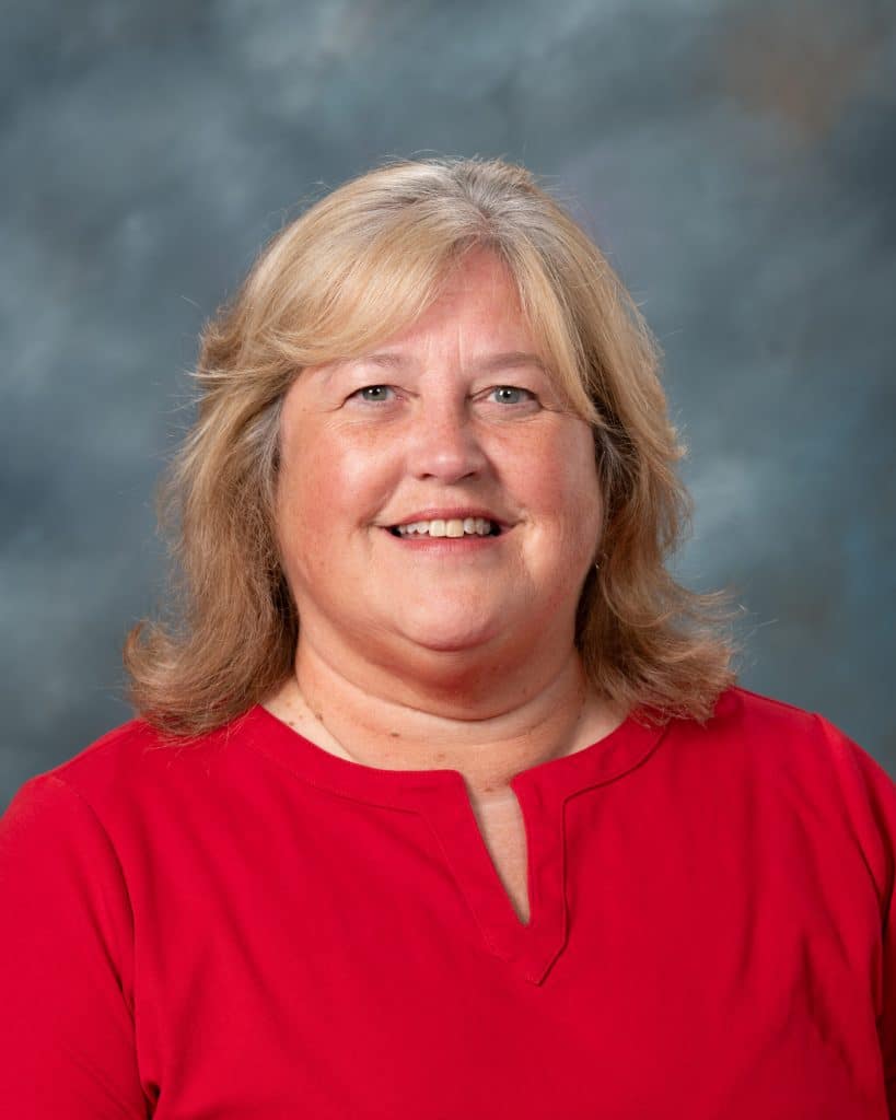 A woman with shoulder-length blonde hair and a friendly smile, wearing a red blouse, poses in front of a gray-blue mottled background.