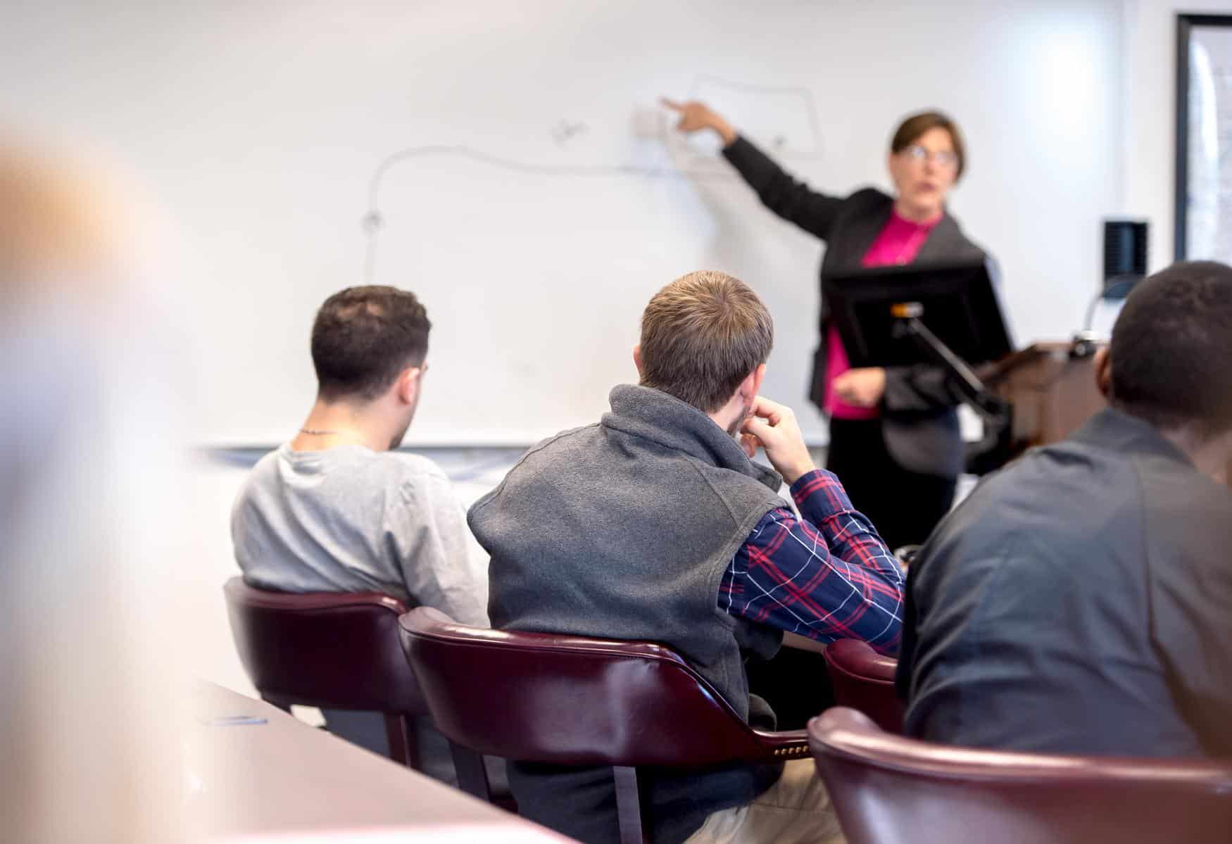 A teacher stands at the front of a classroom, pointing at a whiteboard with a marker. Several students are seated, facing the whiteboard and listening attentively to the lesson.