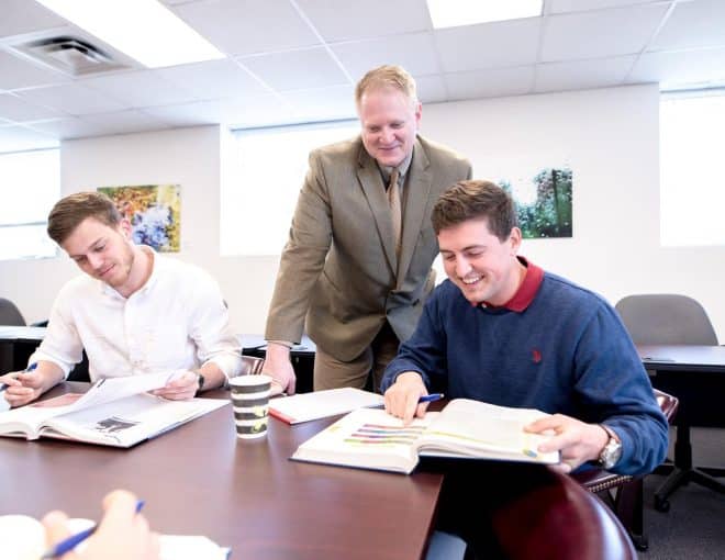A teacher stands and smiles while assisting two male students studying at a table with open books and notebooks in a brightly lit classroom.