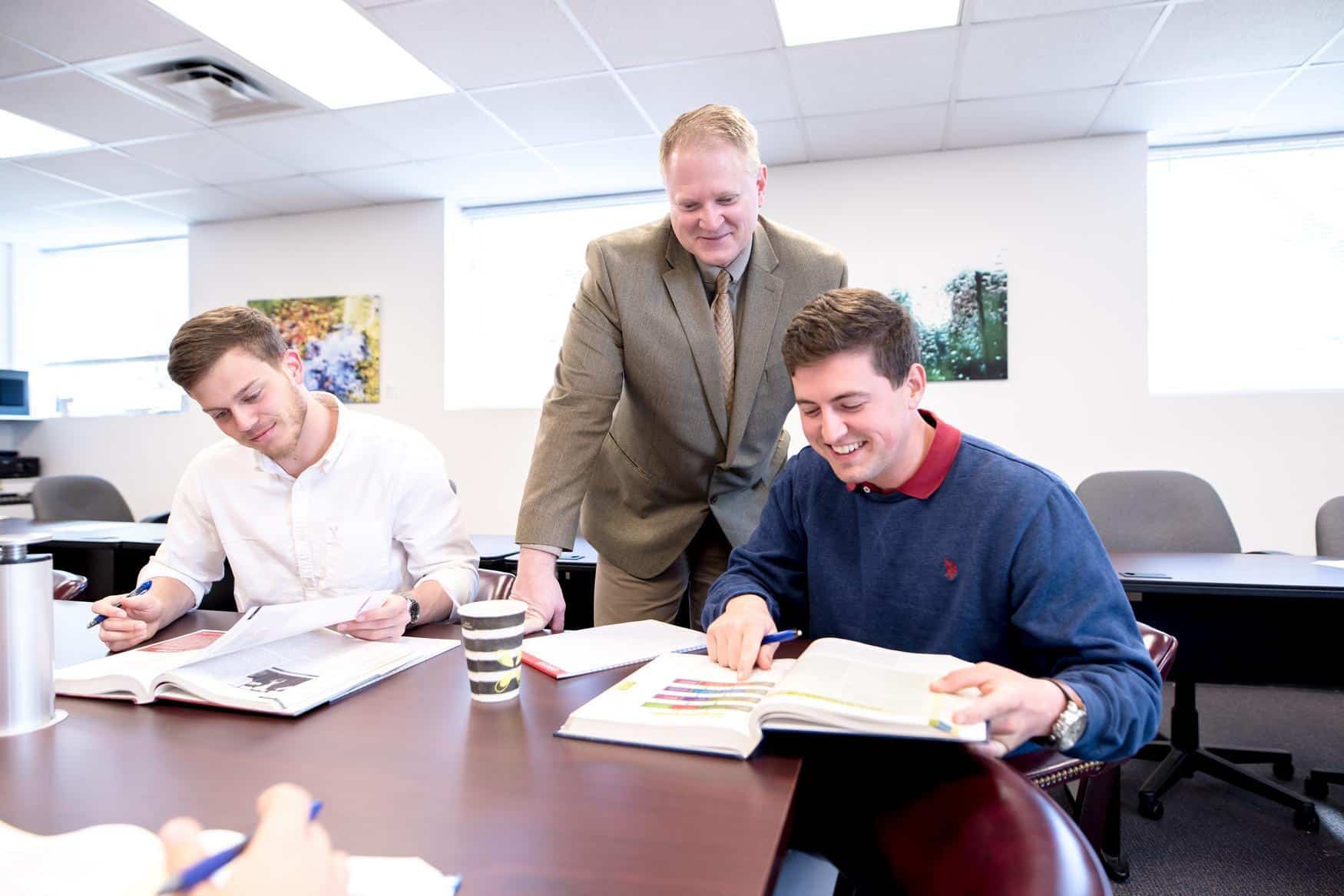 A teacher stands and smiles while assisting two male students studying at a table with open books and notebooks in a brightly lit classroom.
