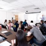 A teacher stands at the whiteboard explaining material to a classroom of students seated at desks, some taking notes and others listening attentively. The classroom is bright with overhead lights.