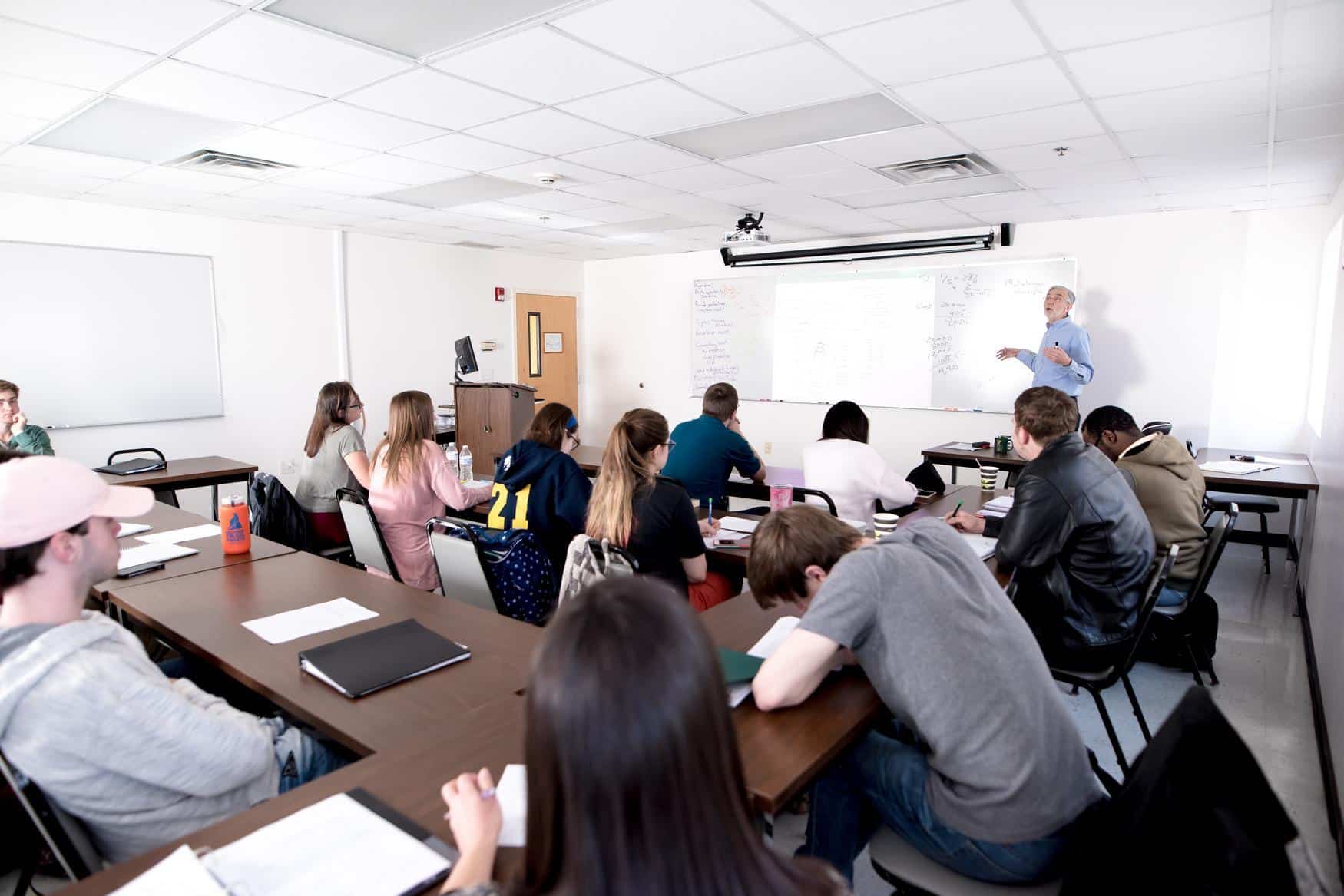 A teacher stands at the whiteboard explaining material to a classroom of students seated at desks, some taking notes and others listening attentively. The classroom is bright with overhead lights.