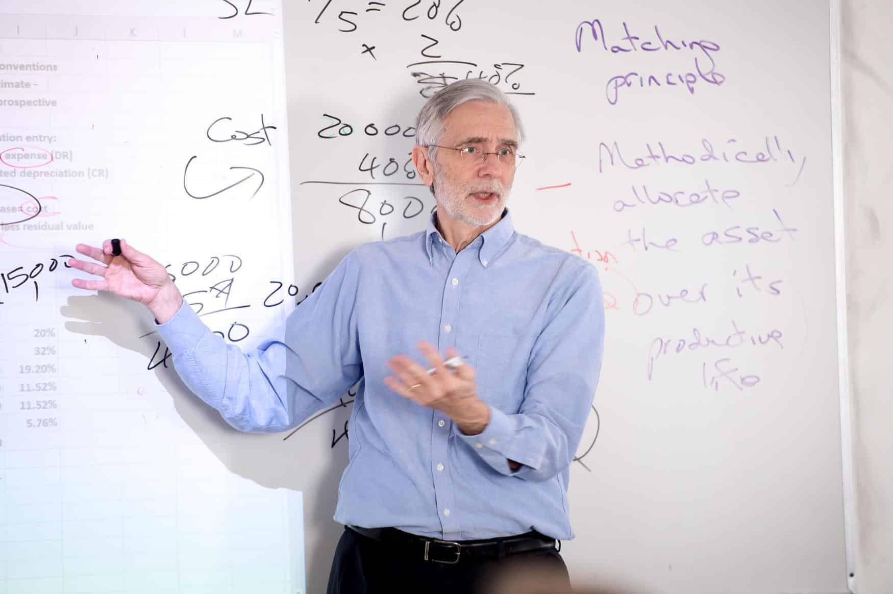 An older man in a blue shirt stands in front of a whiteboard covered with handwritten notes and diagrams, teaching or explaining something while gesturing with his hands.