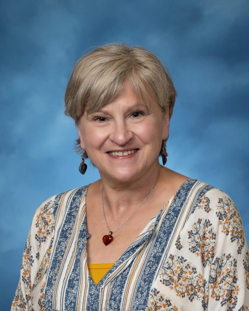 A smiling older woman with short, light brown hair wears a floral blouse, heart-shaped necklace, and matching earrings, posing in front of a blue studio background.