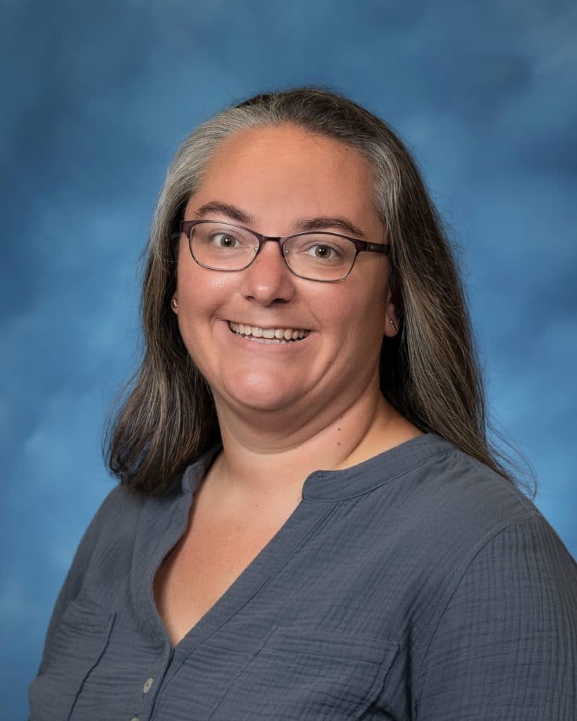 A woman with long brown hair and gray streaks, wearing glasses and a gray blouse, smiles at the camera in front of a blue backdrop.