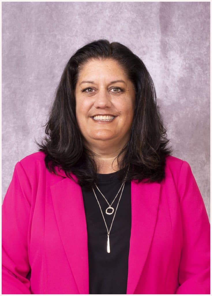 A woman with long dark hair wearing a bright pink blazer over a black top, smiling at the camera. She has layered necklaces and stands against a light gray textured background.