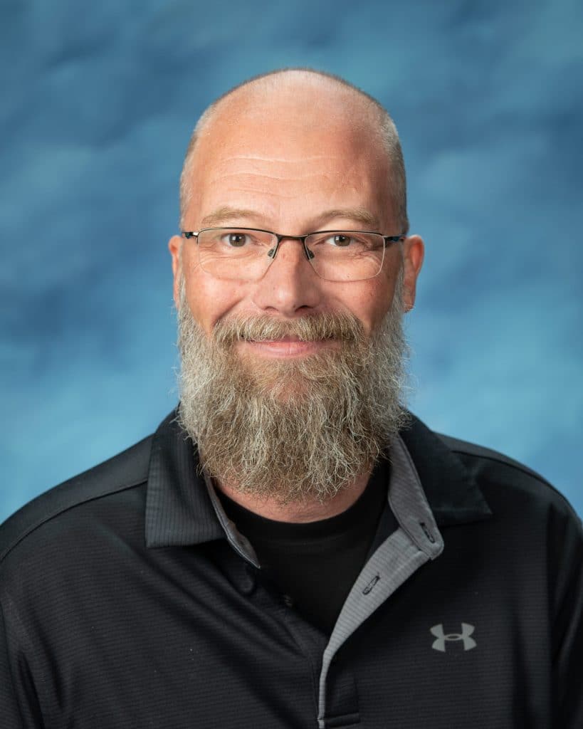 A middle-aged man with a bald head, glasses, and a long gray beard smiles at the camera. He is wearing a black collared Under Armour shirt, and the background is a blue gradient.