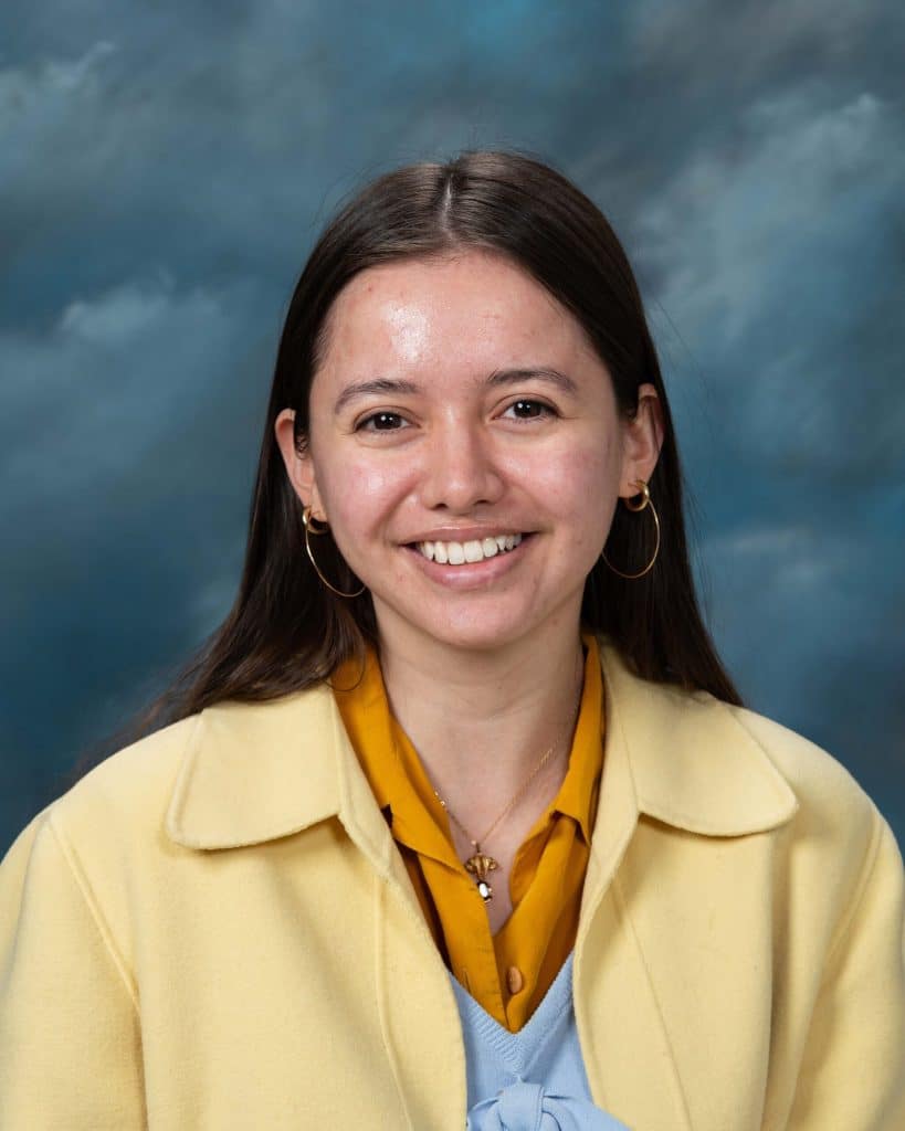 A woman with long brown hair, wearing a yellow coat, gold hoop earrings, and a mustard yellow shirt, smiles in front of a blue and gray cloudy backdrop.