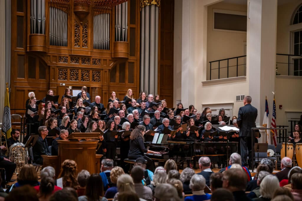 A large choir and orchestra perform on a well-lit stage in a church, directed by a conductor. The audience is seated in pews, and a grand organ is visible in the background.