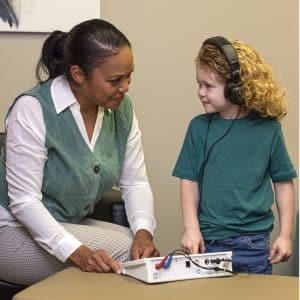 A woman conducts a hearing test for a young child wearing headphones. The woman adjusts controls on an audiometer while smiling at the child, who is looking back and smiling.