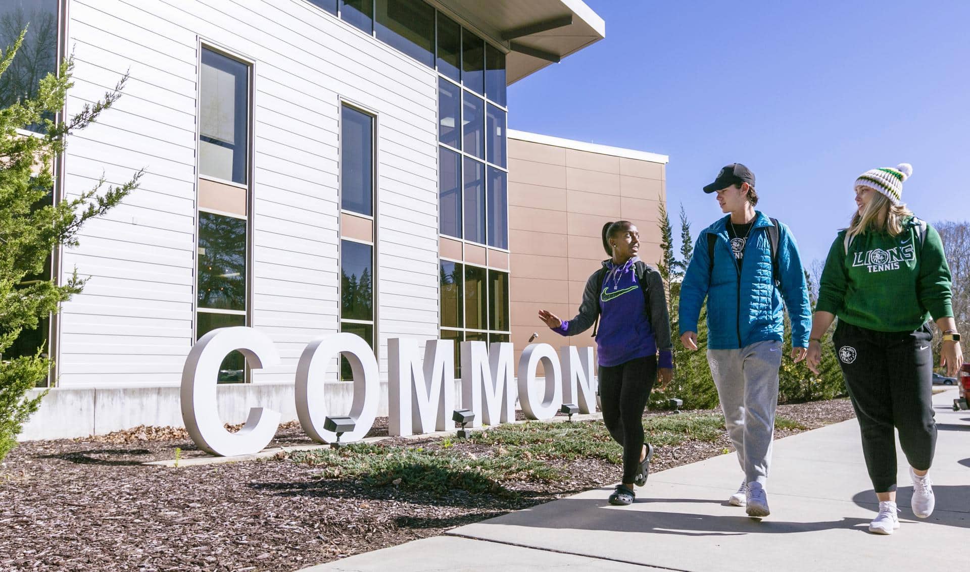 Three people walk and talk on a sidewalk outside a modern building, passing large white letters spelling COMMON. The sky is clear and blue, and everyone is dressed casually for cool weather.