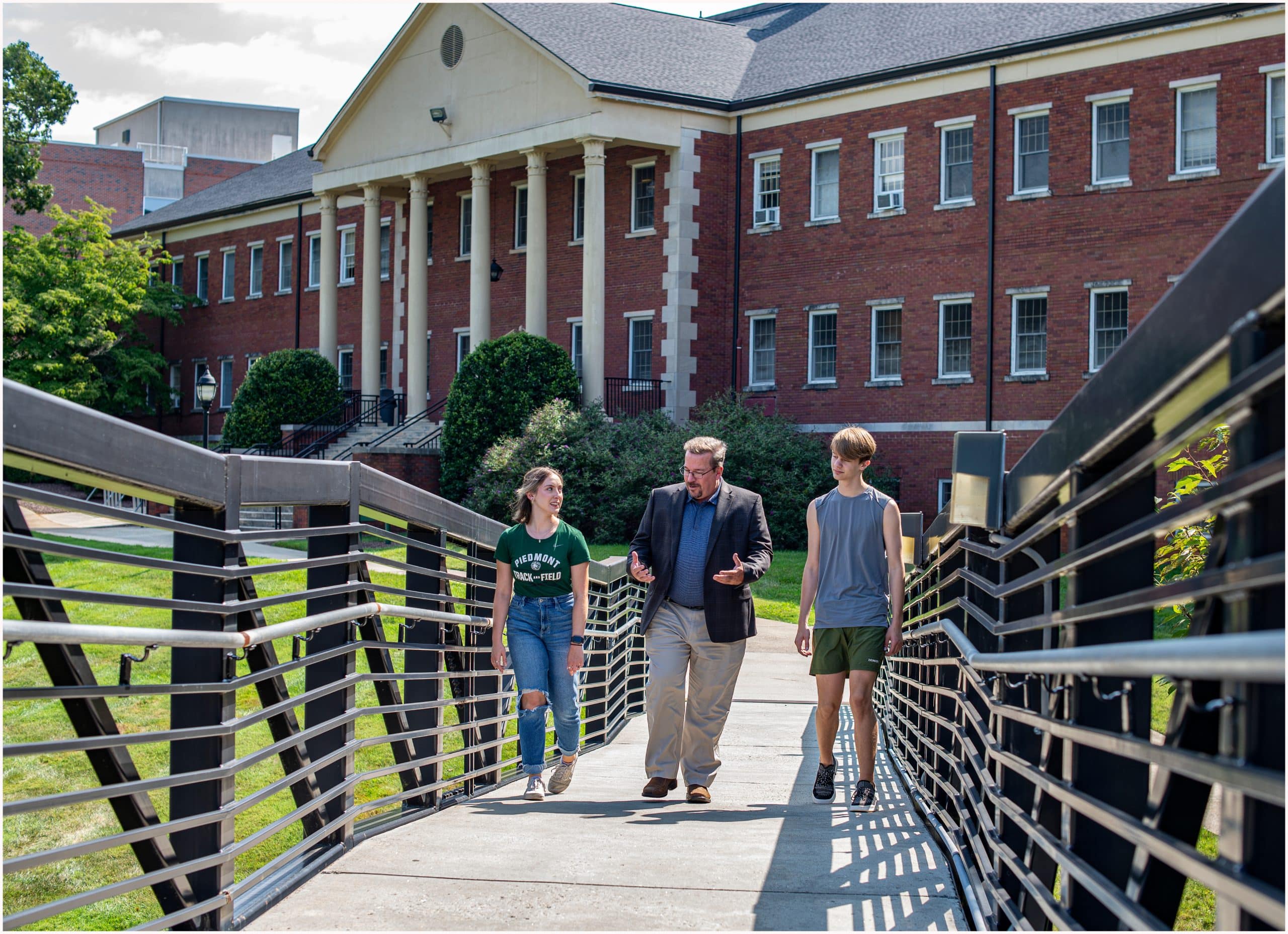 A man and two students walk across a pedestrian bridge toward a red brick academic building with white columns on a sunny day at Piedmont University. The man is talking and gesturing while the students listen.
