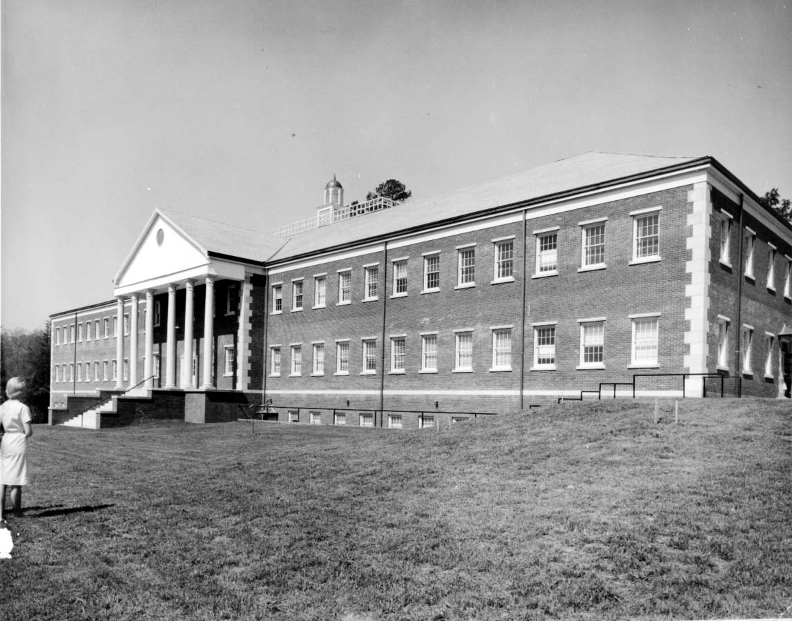A black-and-white photo of a large brick building with two stories, tall columns at the entrance, and many windows. A person stands on the grass in the foreground, looking toward the building.