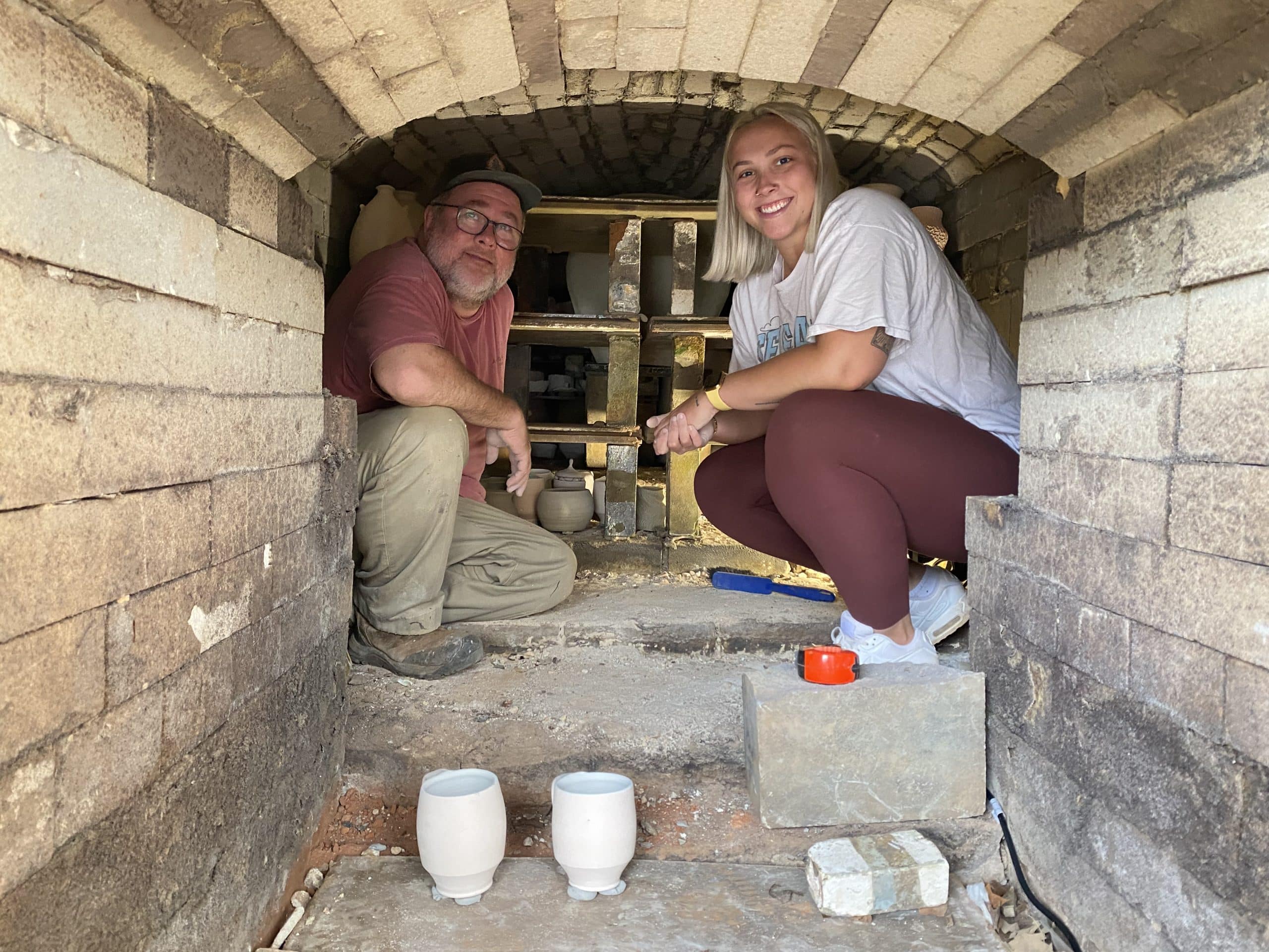 Two people squatting inside of a kiln, in front of rows of ceramics