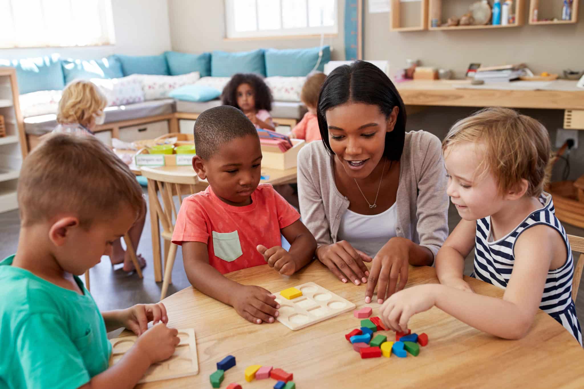 Woman with a group of young children at a table. The children are playing with a wooden puzzle and listening to the woman.