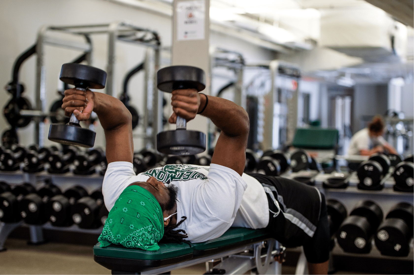 A young man in a gym, wearing a green bandana laying on a weight bench lifting free weights.