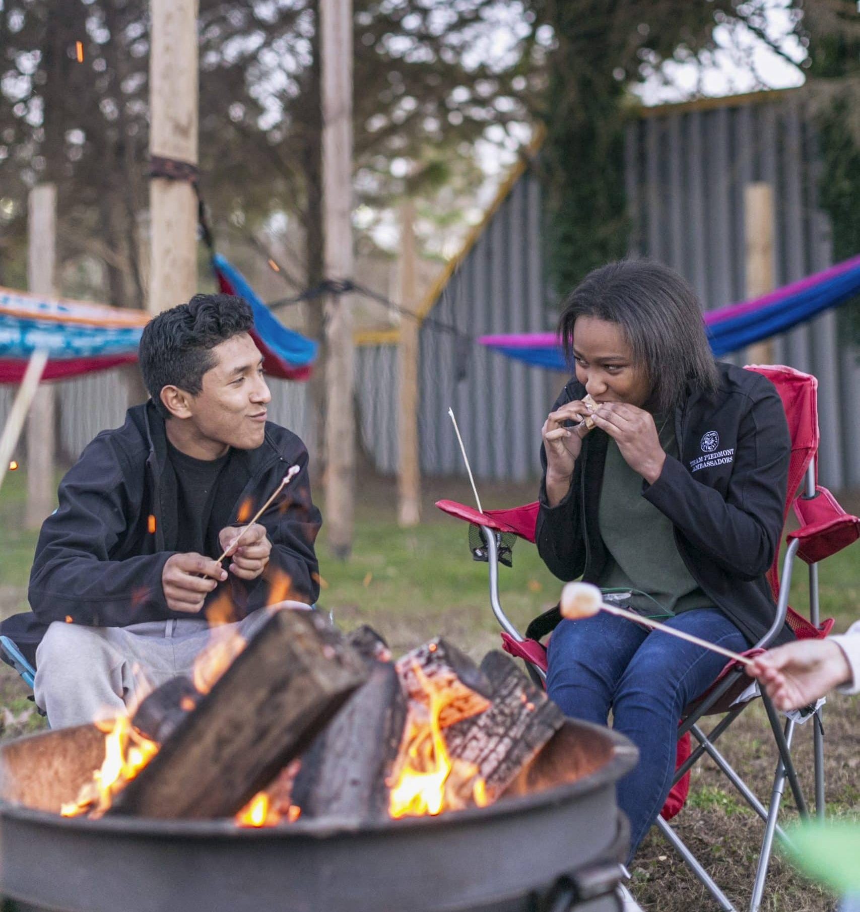 Students in camping chairs in front of a fire. They are smiling and eating smores.