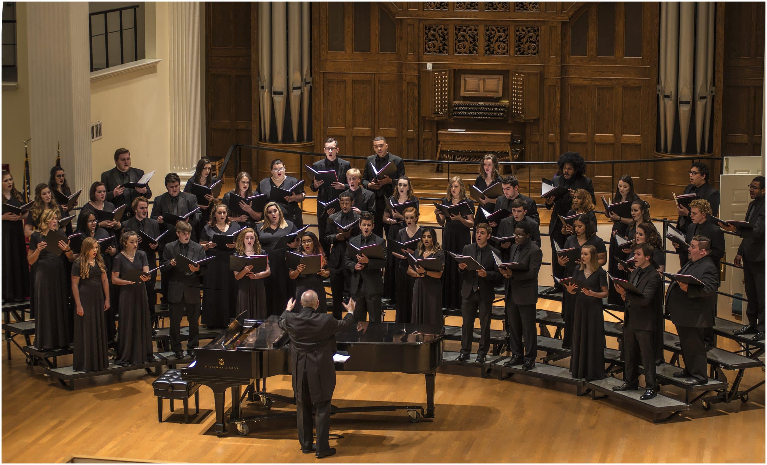 Choral group in black clothing performing. A conductor leads them, standing in front of a black piano.