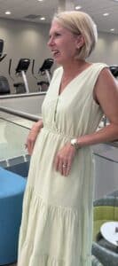 A smiling woman with short blonde hair wearing a sleeveless light green dress stands indoors near a railing. Exercise machines are visible in the background.
