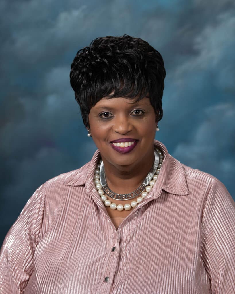 A woman with short black hair, wearing a shiny pink blouse and layered pearl necklaces, smiles in front of a blue and gray mottled background.