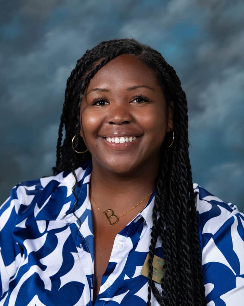A woman with long, twisted hair and hoop earrings smiles at the camera. She wears a blue and white patterned blouse and a gold necklace with the letter B against a blurred blue-gray background.