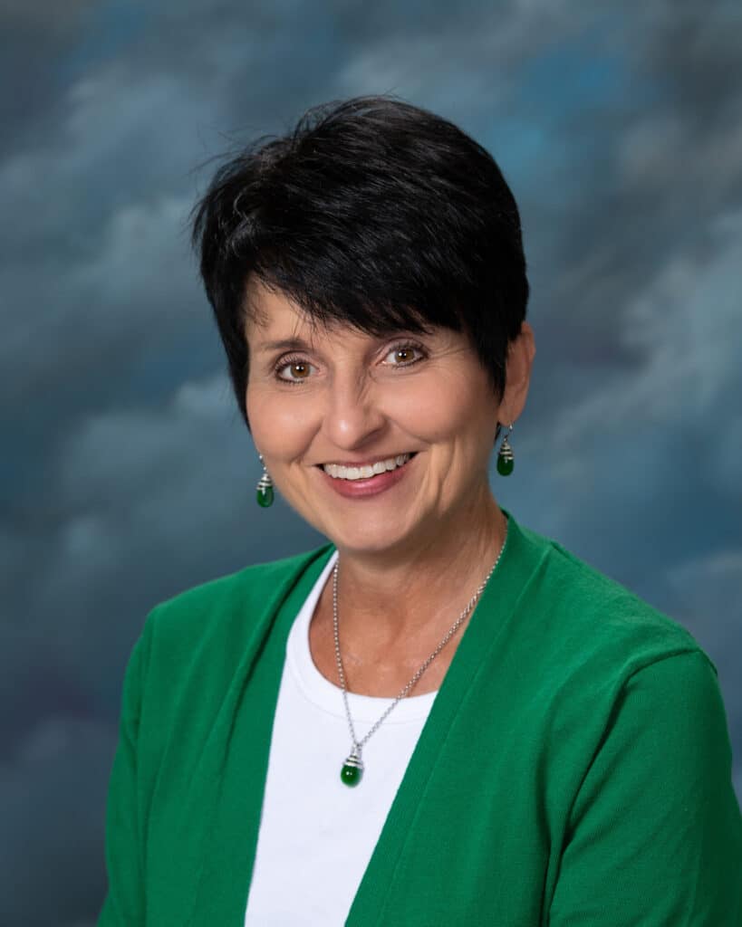 A woman with short dark hair, wearing a green cardigan over a white top, matching green earrings and necklace, smiles at the camera against a cloudy blue background.