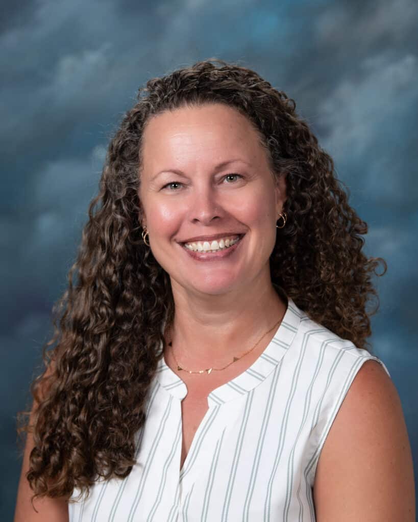 A woman with long curly brown hair smiles at the camera. She is wearing a sleeveless white blouse with thin vertical stripes and gold hoop earrings, posed in front of a cloudy blue and gray backdrop.