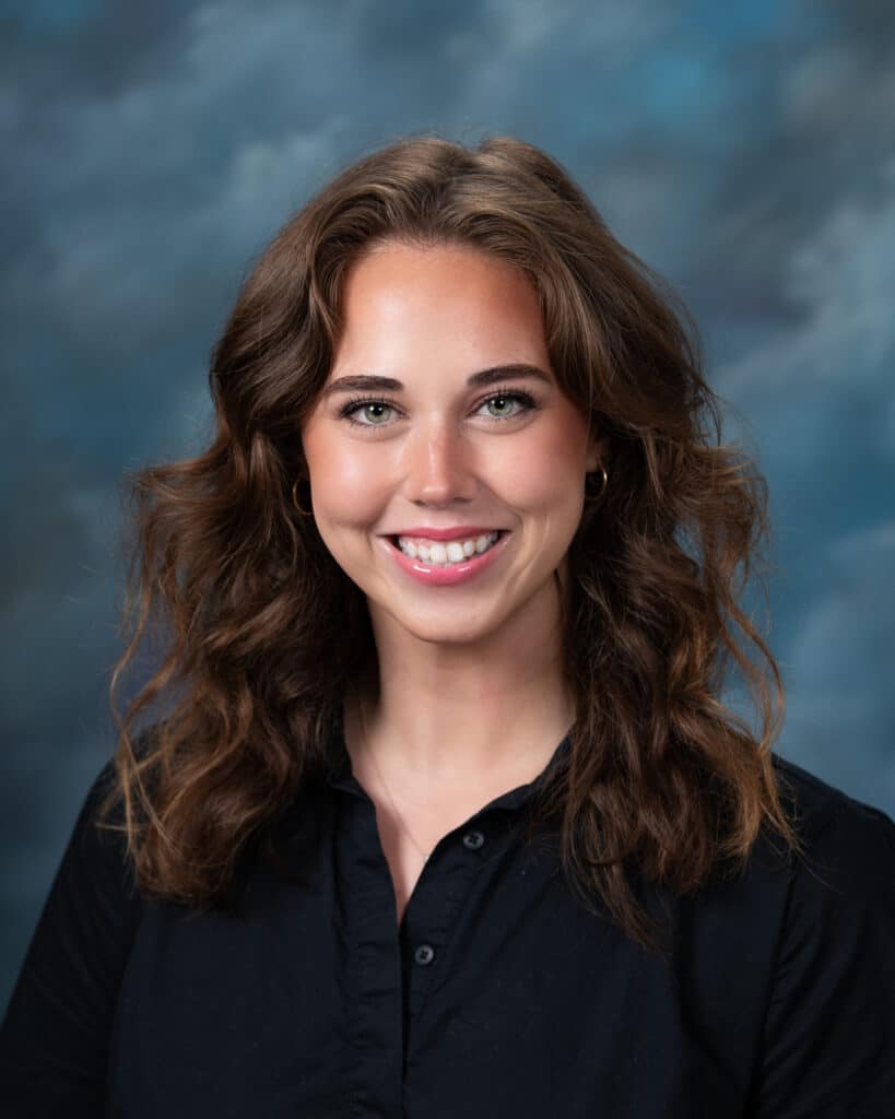 A young woman with wavy brown hair, wearing a black collared shirt, smiles at the camera against a blue and gray studio backdrop.