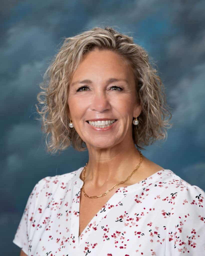 A woman with short, curly blonde hair is smiling. She wears a white blouse with a pink floral pattern, gold necklace, and pearl earrings. The background is a soft, blurred blue and gray.