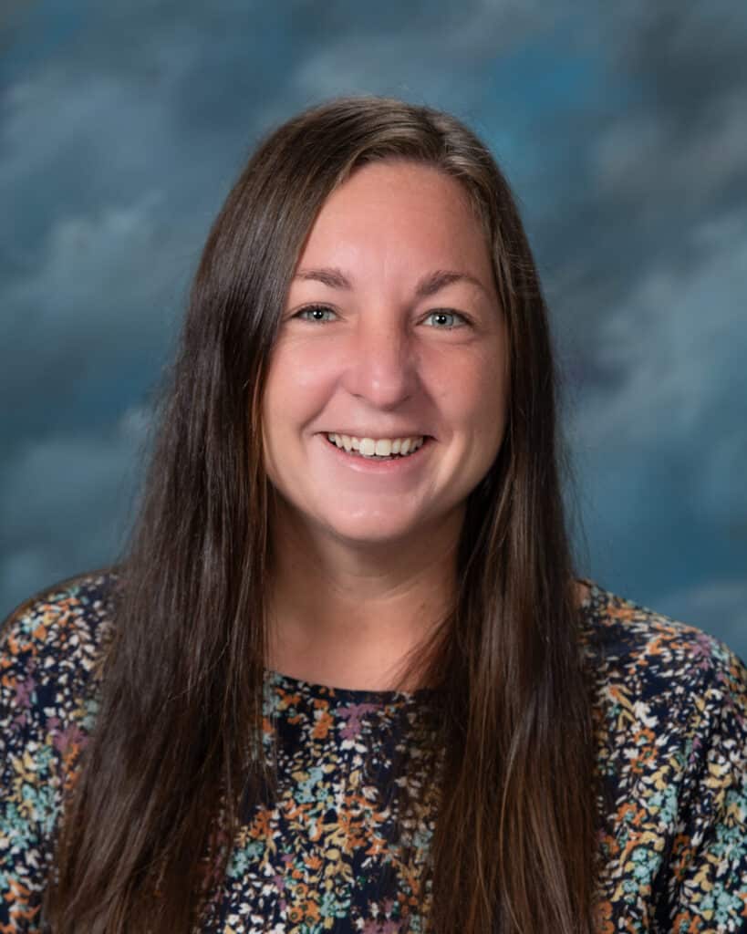 A woman with long brown hair and green eyes smiles at the camera. She is wearing a floral patterned top and is posed in front of a blue and gray cloudy background.